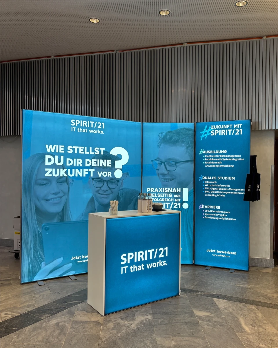 Zu sehen ist ein moderner Messestand mit vier Elementen: dem größten Element links mit den Schriftzug "Wie stellst du dir eine Zukunft vor?", links daneben zwei kleinere Elemente mit den Ausbildungs- und Studienangeboten bei SPIRIT/21. Im Vordergrund steht noch ein SPIRIT/21-gebrandeter Tresen | SPIRIT/21