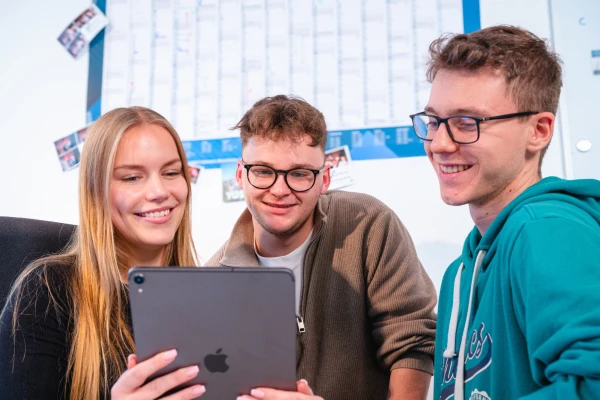 Three young people are looking at an iPad: a blonde white woman on the left and two dark blonde men with glasses in the middle and on the right. In the background you can see a large wall calendar | SPIRIT/21