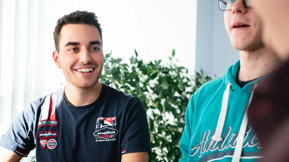 Ein junger Mann mit dunklen Haaren und einem dunkelblauen T-Shirt mit Aufdrucken lächelt im Gespräch mit anderen Auszubildenen | SPIRIT/21