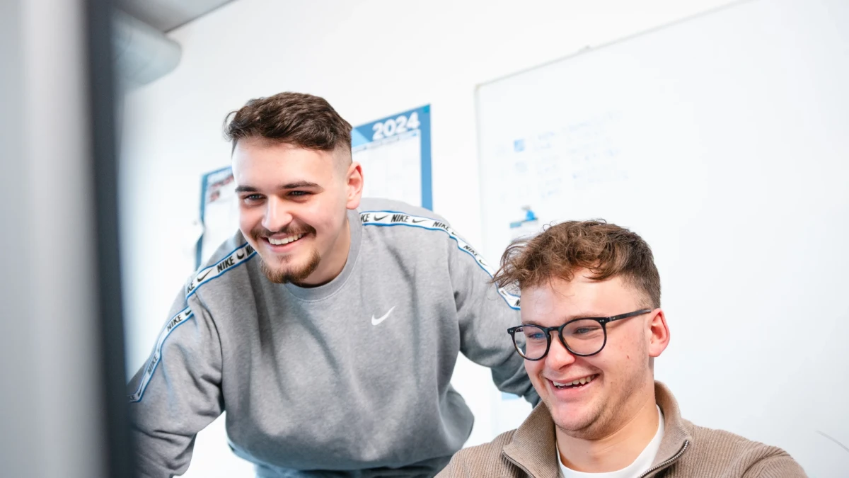 Ein junger Mann lehnt sich von hinten zum Arbeitsplatz mit Monitor eines anderen jungen Mannes mit Brille, der am Arbeitsplatz sitzt. | SPIRIT/21