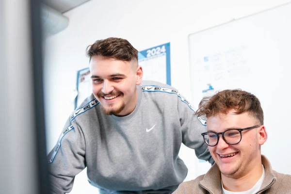 Ein junger Mann lehnt sich von hinten zum Arbeitsplatz mit Monitor eines anderen jungen Mannes mit Brille, der am Arbeitsplatz sitzt. | SPIRIT/21