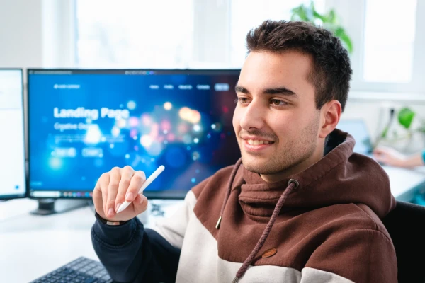 Ein junger Mann mit dunklen Haaren sitzt auf einem Bürostuhl. In der Hand hält er einen Stit für ein Tablet. Im Hintergrund sieht man Monnitore. | SPIRIT/21