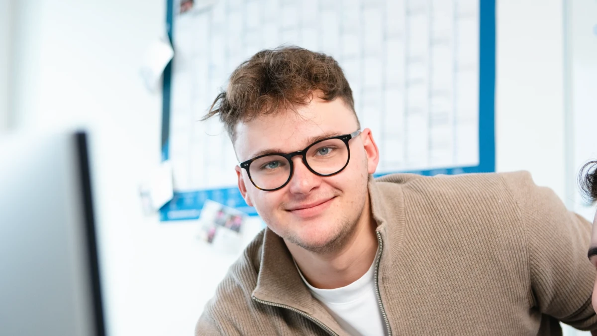 Ein junger Mann mit Brille lehnt sich über einen Arbeitsplatz im Büro. Im Hintergrund ist ein Wandkalender sichtbar. | SPIRIT/21