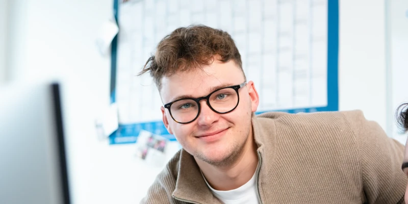 Ein junger Mann mit Brille lehnt sich über einen Arbeitsplatz im Büro. Im Hintergrund ist ein Wandkalender sichtbar. | SPIRIT/21