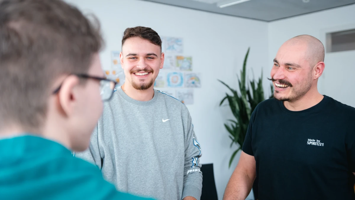 Über dir Schulter Blick eines Kollegen auf zwei weitere Kollegen, die sich in einem Gespräch in einem Besprechungsraum befinden | SPIRIT/21