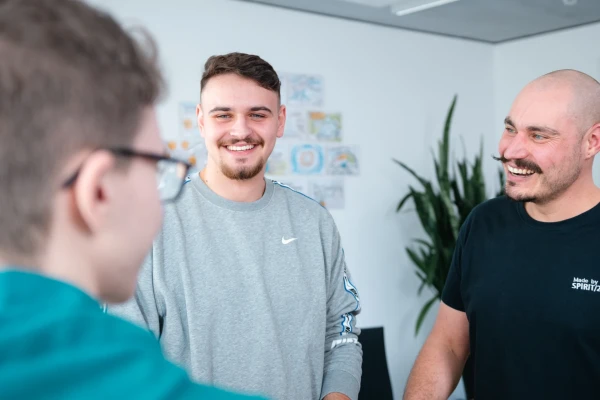 Über dir Schulter Blick eines Kollegen auf zwei weitere Kollegen, die sich in einem Gespräch in einem Besprechungsraum befinden | SPIRIT/21