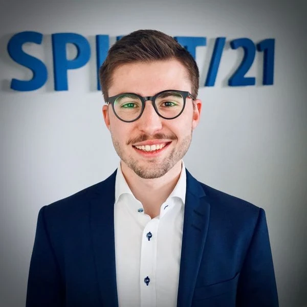 Portraitfoto eines jungen Mannes mit dunklen Haaren, dunklen Augen und Brille, der in die Kamera lächelnt. Im Hintergrund ist das SPIRIT/21-Logo | SPIRIT/21