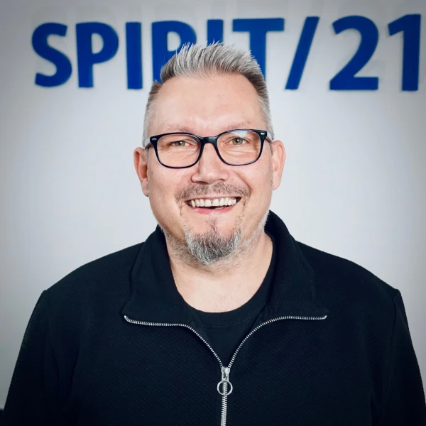 Portraitfoto eines mittelalten Mannes mit gräulichen Haaren und einer Brille, der herzlich in die Kamera lacht. Im Hintergrund ist das SPIRIT/21-Logo | SPIRIT/21