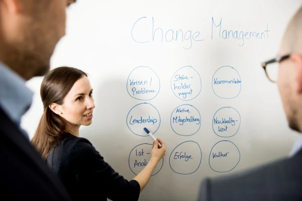 Eine Frau steht vor einem Whiteboard und zeigt mit einem Marker auf ein Kommentar, welcher auf diesem steht. Zwei Männer hören ihr zu | SPIRIT/21