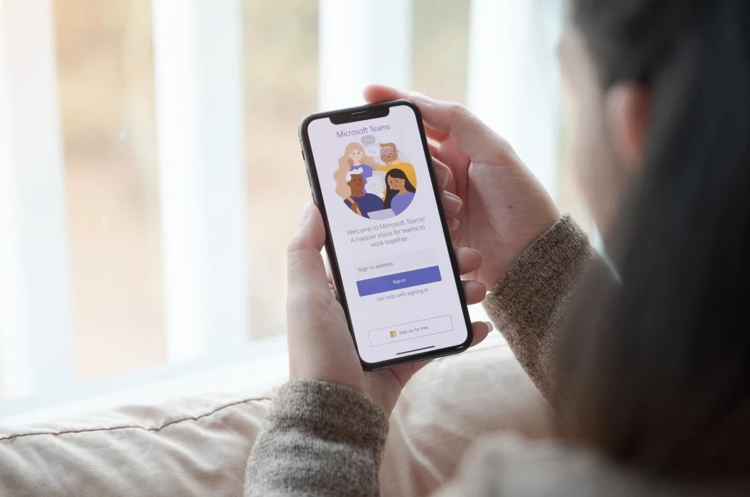 Über die Schulter Blick einer Frau mit dunklen Haaren, welche ein Smartphone hält. Auf dem Display ist die Anmeldemaske für Microsoft Teams zu sehen. | SPIRIT/21