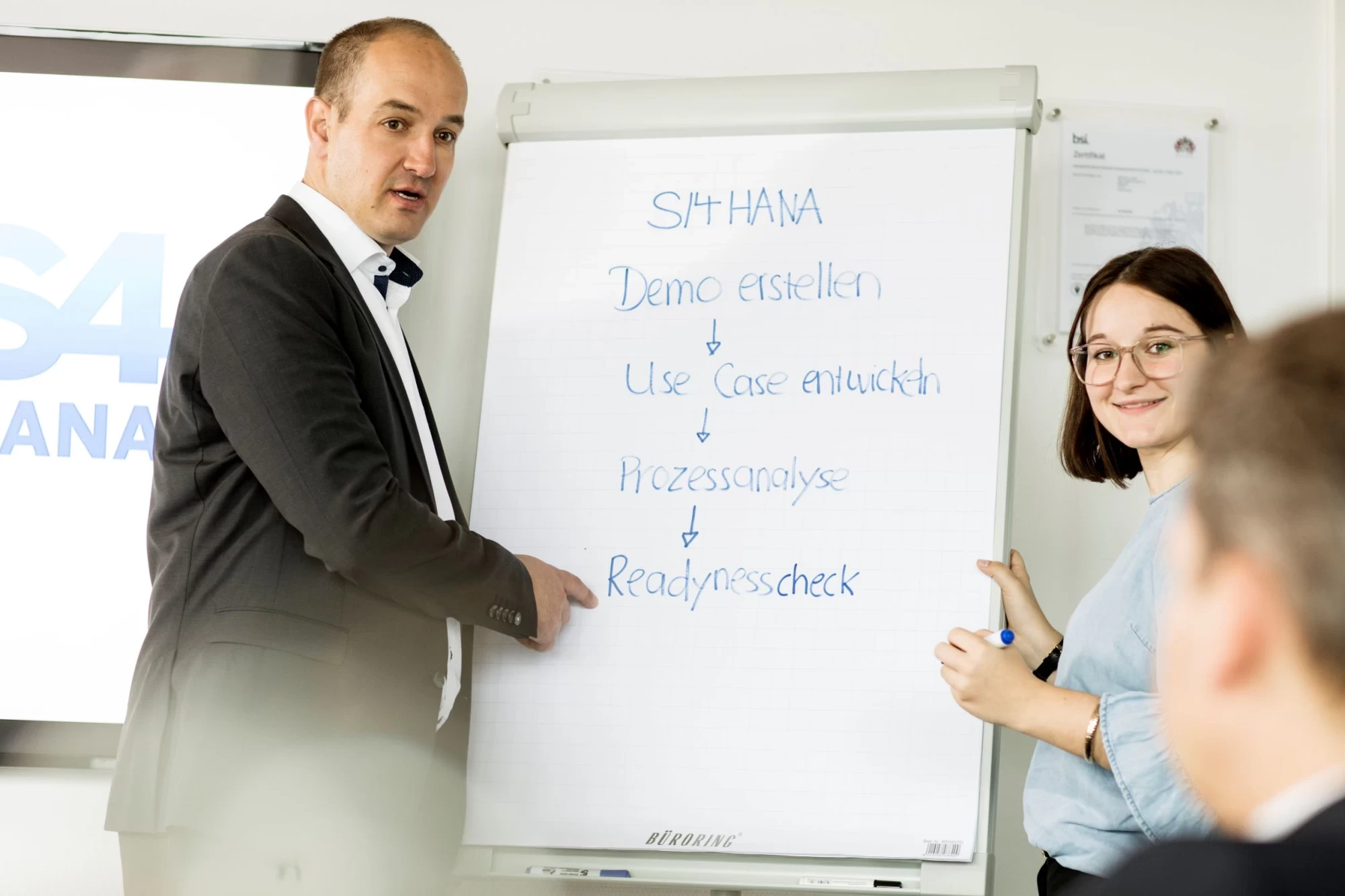 Ein Mann (links) und eine Frau (rechts) stehen vor einem Whiteboard und präsentieren in geschäftlichen Bereich das Thema SAP S/4HANA | SPIRIT/21