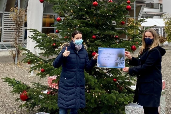 Übergabe des Spendenscheins: Zwei Frauen stehen vor einem geschmückten Weihnachtsbaum und haben einen großen Scheck in der Hand | SPIRIT/21
