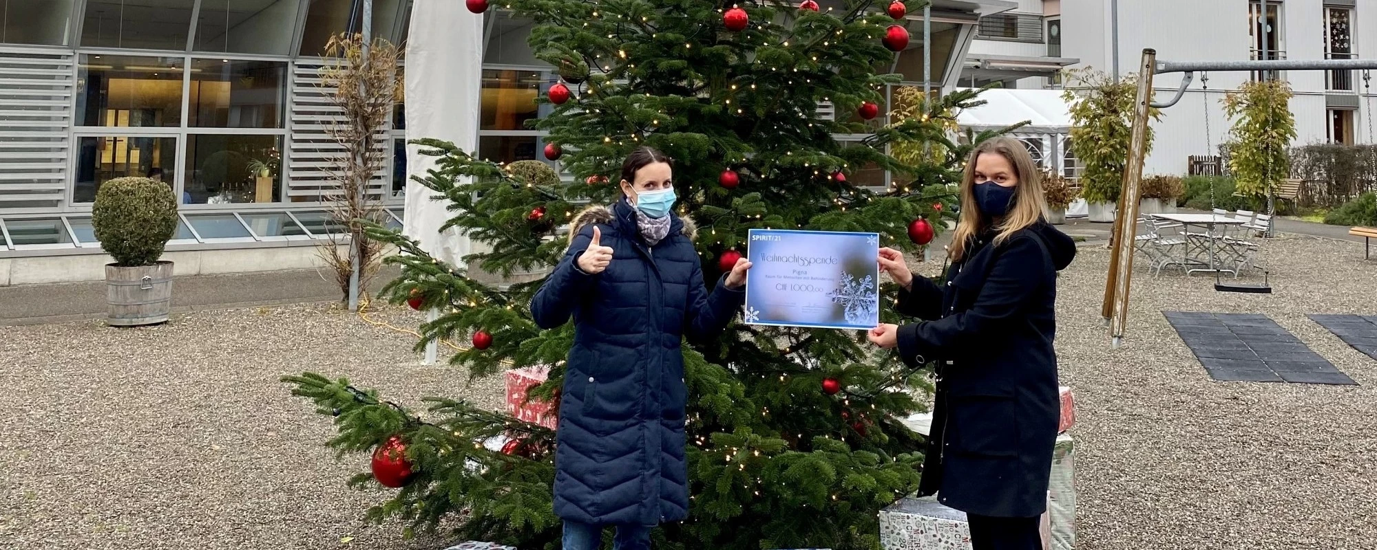 Übergabe des Spendenscheins: Zwei Frauen stehen vor einem geschmückten Weihnachtsbaum und haben einen großen Scheck in der Hand | SPIRIT/21