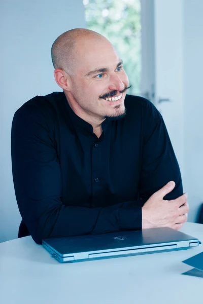 Seitenansicht einer Person mit rasiertem Kopf und Bart, l&auml;ssige Sitzhaltung, heller Innenraum, runder Bildausschnitt | SPIRIT/21